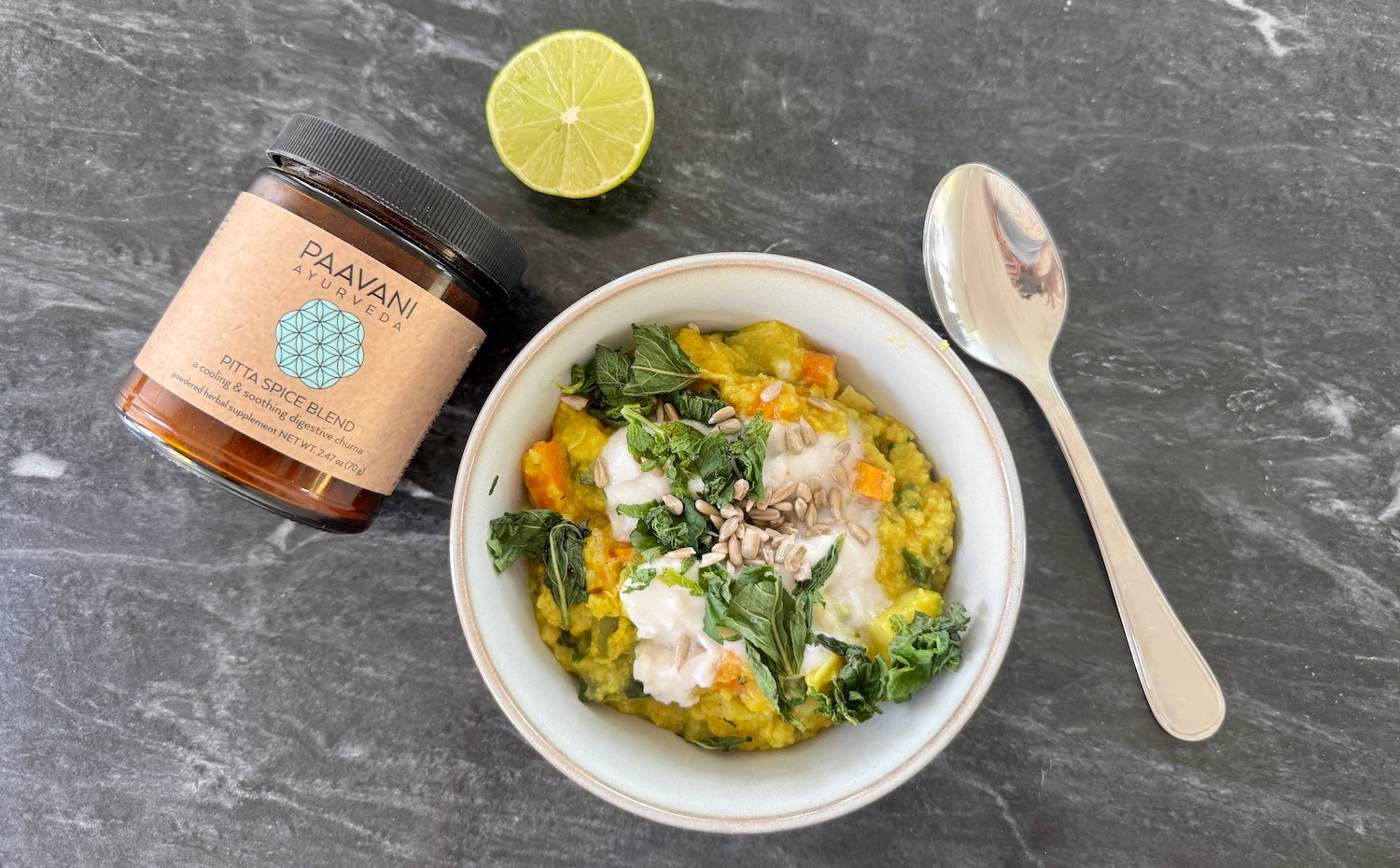 A light, summery kitchari served in a white ceramic bowl, garnished with fresh herbs, surrounded by coconut, lime & PAAVANI Pitta Spice Blend.