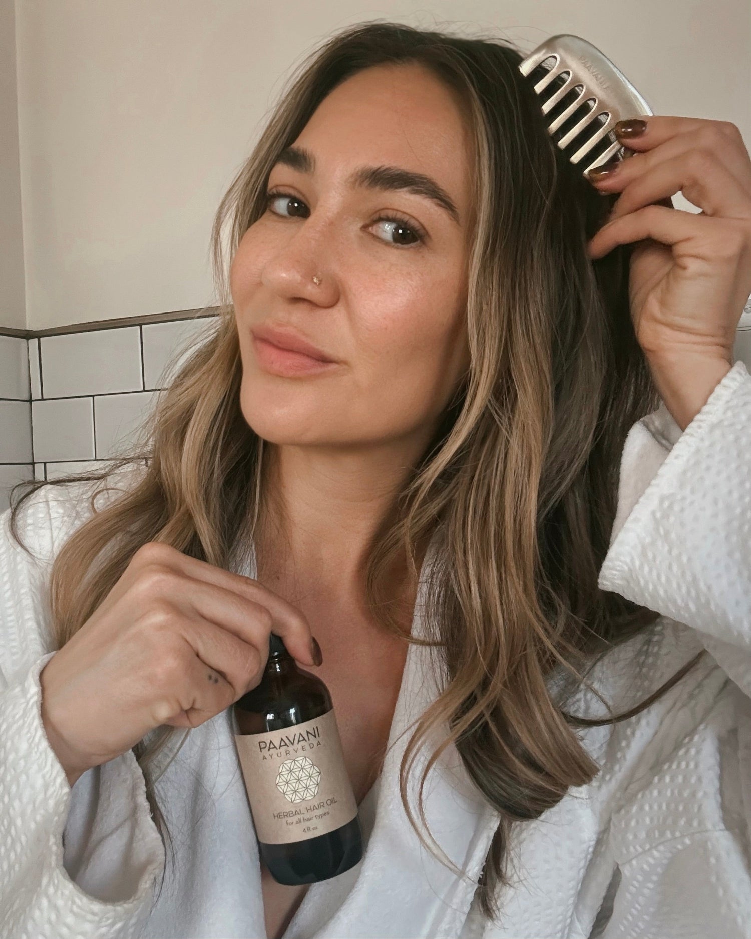 Woman in a white robe holding a bottle of hair oil and a kansa comb, with a tiled wall background.