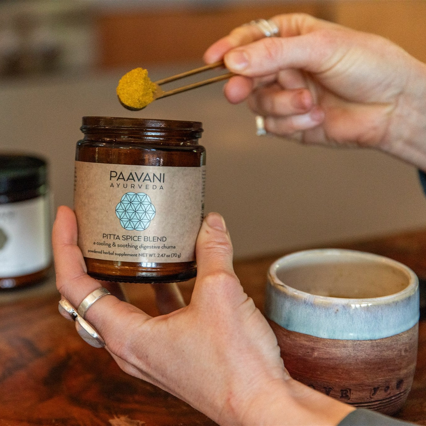 Person holding a jar of Paavani Ayurveda spices with a spoonful above it on a wooden table.