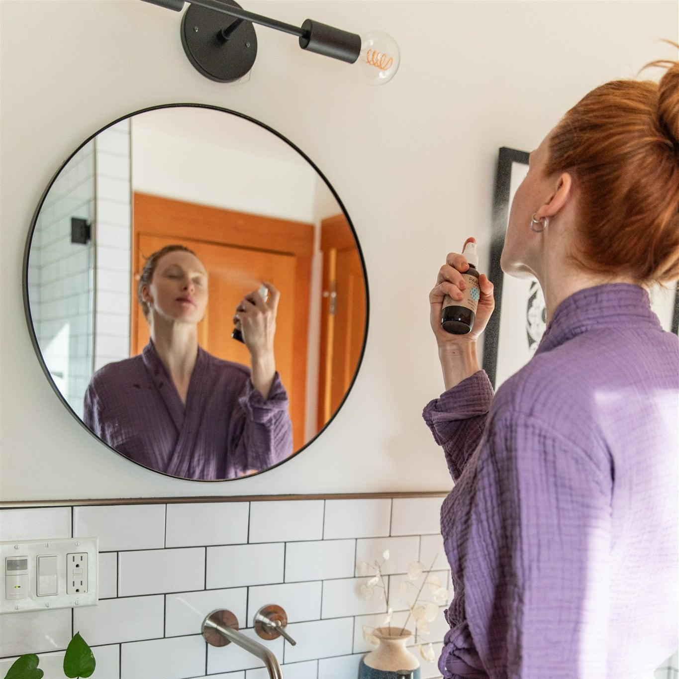 Woman applying PAAVANI Facial Toner in front of a round mirror in a bathroom.
