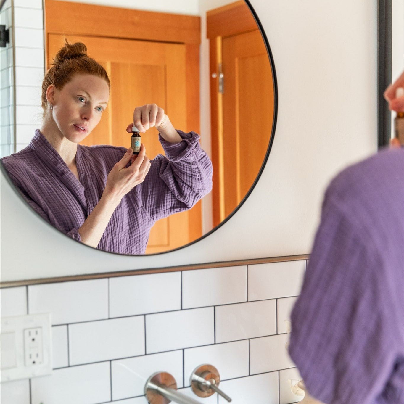 Woman in a purple robe looking at herself about to apply her PAAVANI Facial Serum in a round mirror in a bathroom.
