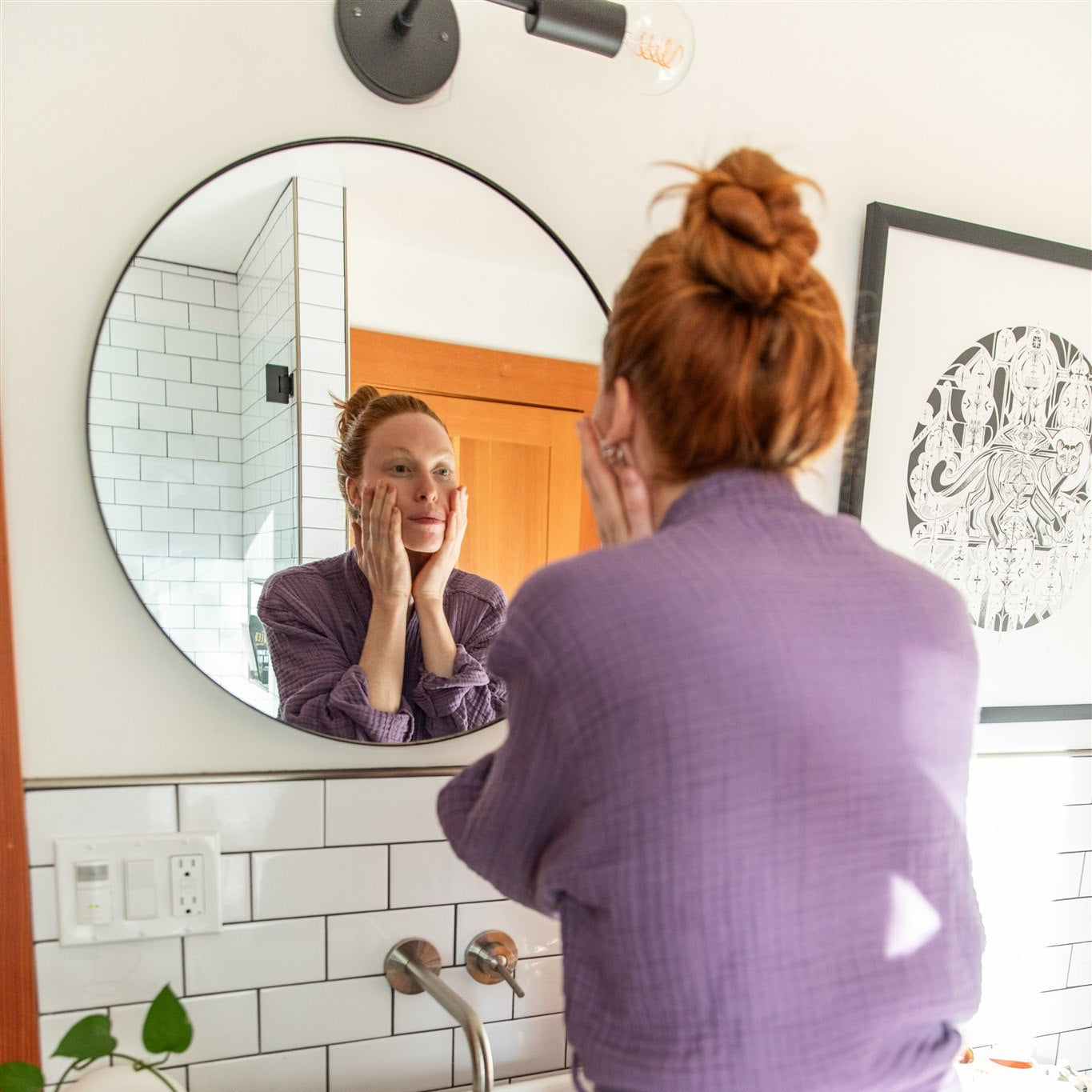 Woman in a purple robe applying PAAVANI Serum to her face in front of a bathroom mirror.