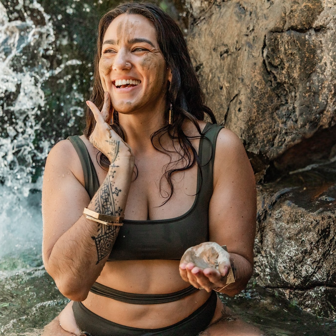 Woman in a swimsuit sitting by a waterfall with rocks and water splashes in the background, trying PAAVANI Clarify Skincare Sampe set