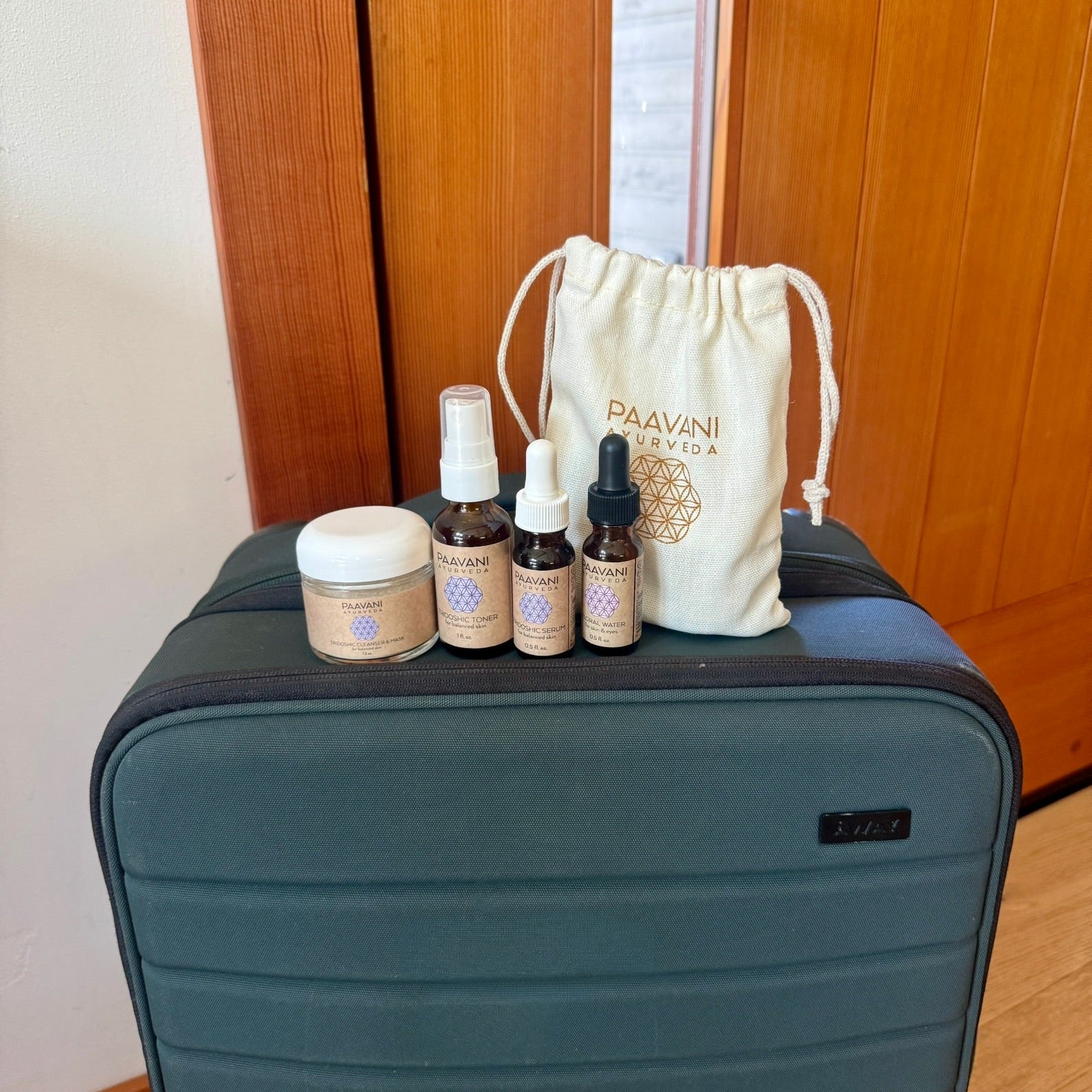 Luggage with PAAVANI Travel size skincare products and a drawstring bag on a wooden floor.