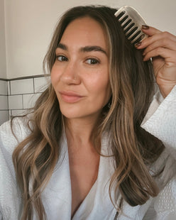 Woman massaging her scalp with a kansa comb in a bathroom wearing a white robe.