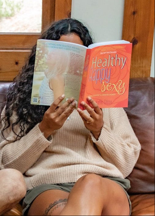 Woman sitting on a couch reading a book with a window in the background.