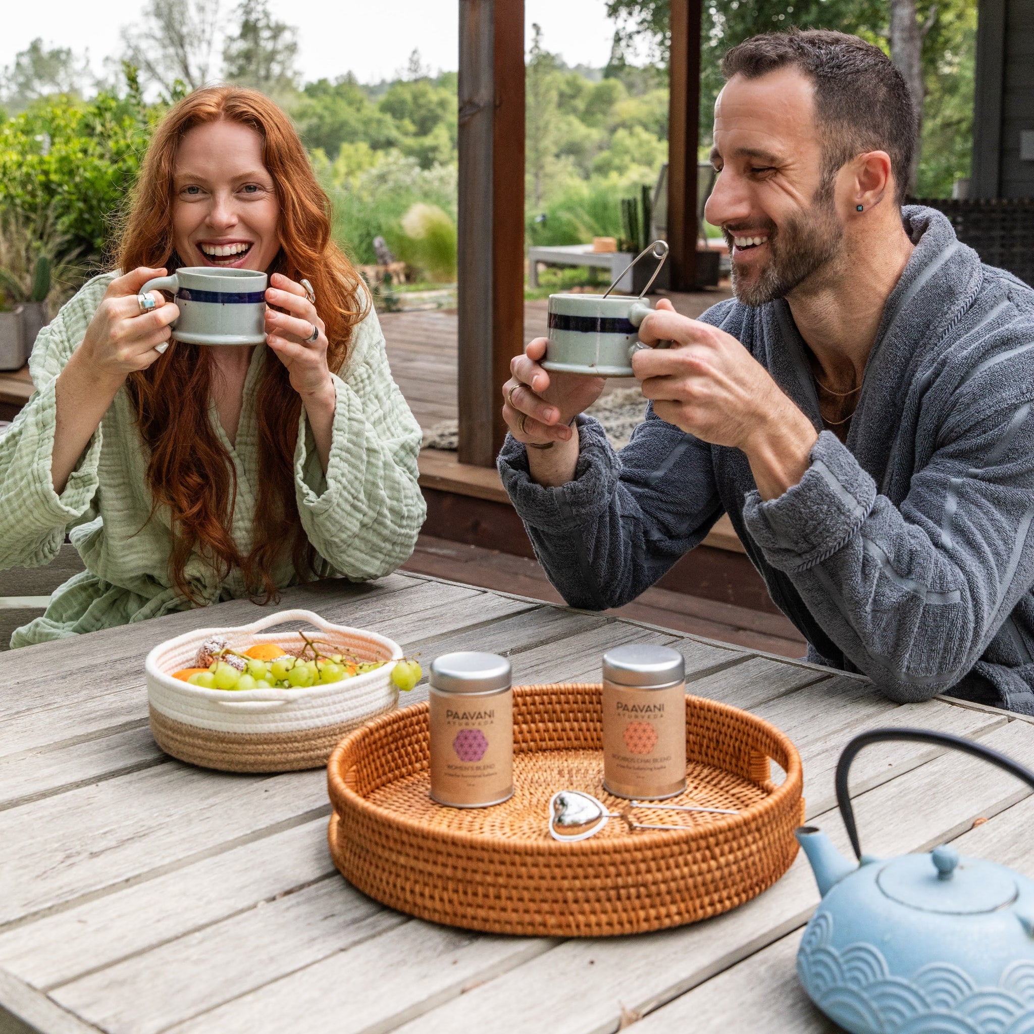 Man and woman enjoying tea outdoors on a wooden deck with a scenic background.