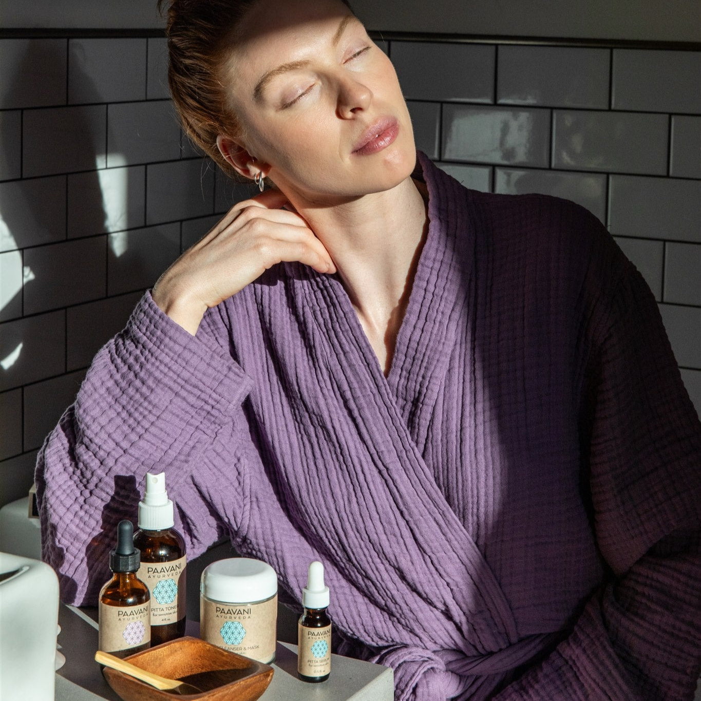 Woman in a purple robe sitting in a bathroom with Paavani Skincare ritual next to her glowing skin.