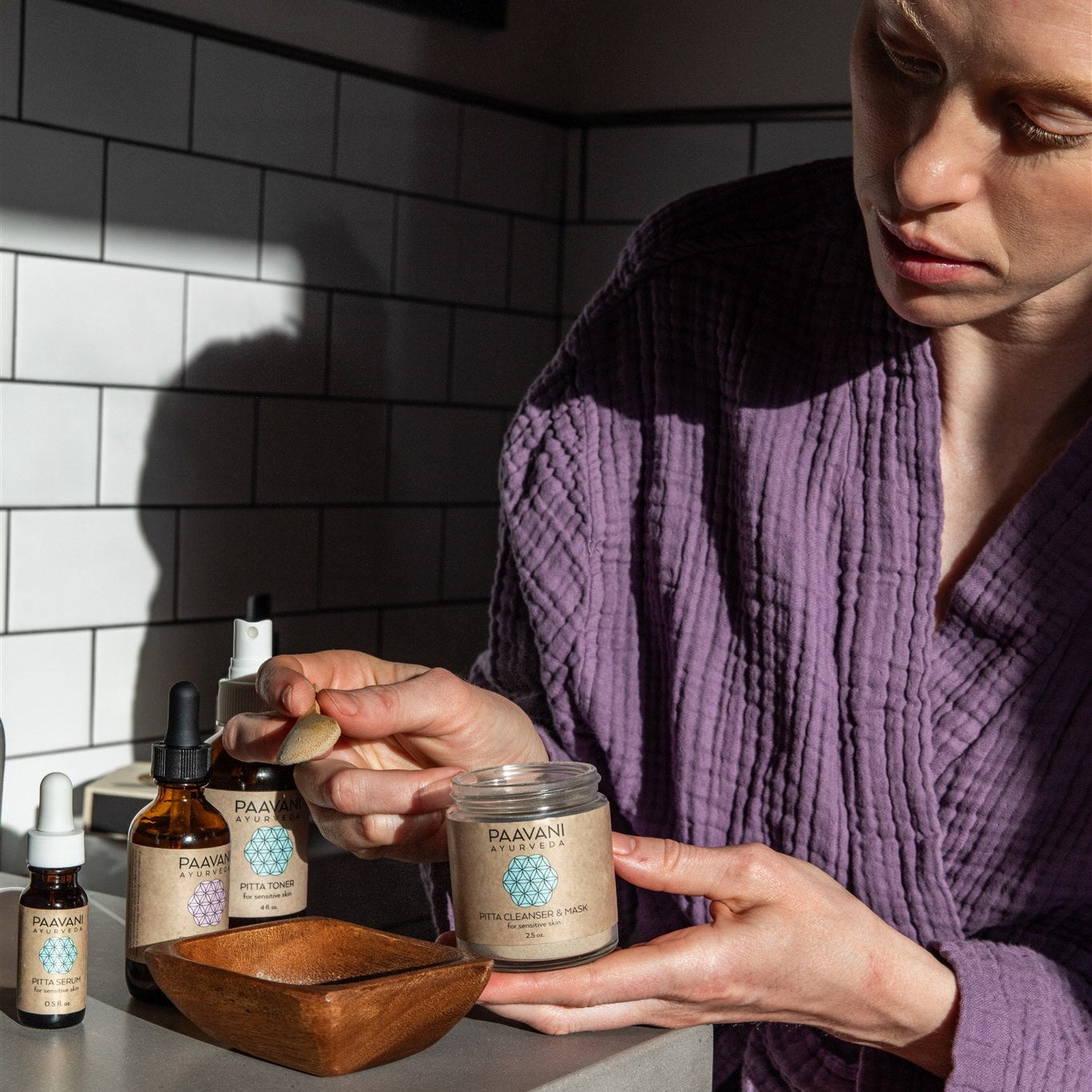 Person in a purple robe preparing a PAAVANI Cleanser & Mask in a bathroom setting.