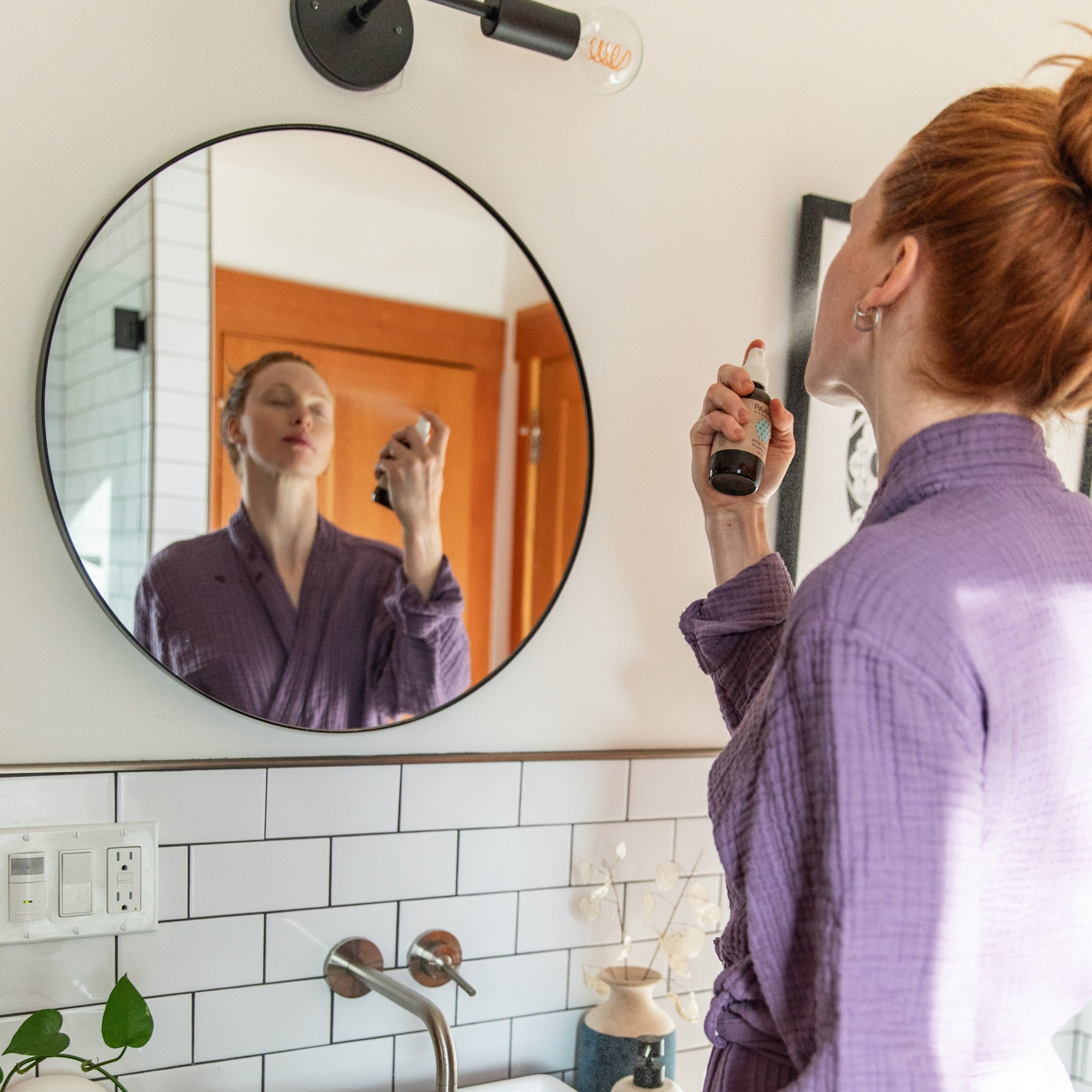 Woman applying makeup in front of a round mirror in a bathroom.